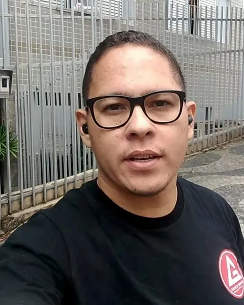 Retrato de Lennildo Bogea, de óculos e camiseta preta, ao ar livre, em frente a um condomínio